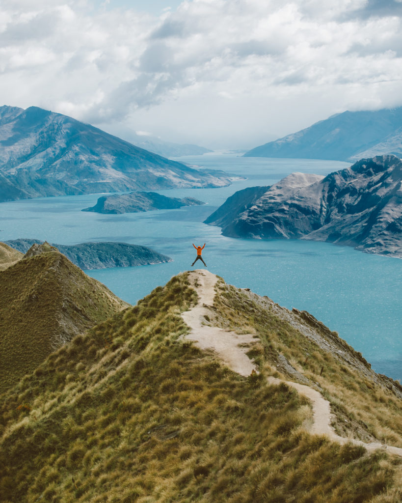 New Zealand Travel Guide | Person jumping in the air at the top of Roy's Peak hike with mountains peeking from blue water in background. 