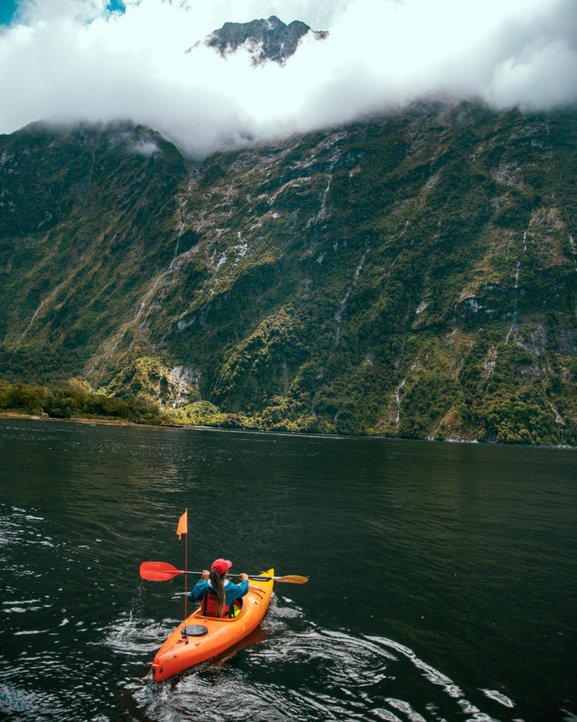 New Zealand Travel Guide | Enormous mossy mountains stand tall amidst mysterious billows of fog as a New Zealand traveler takes a break from paddling small orange canoe, to take in the wonder of Milford Sound.
