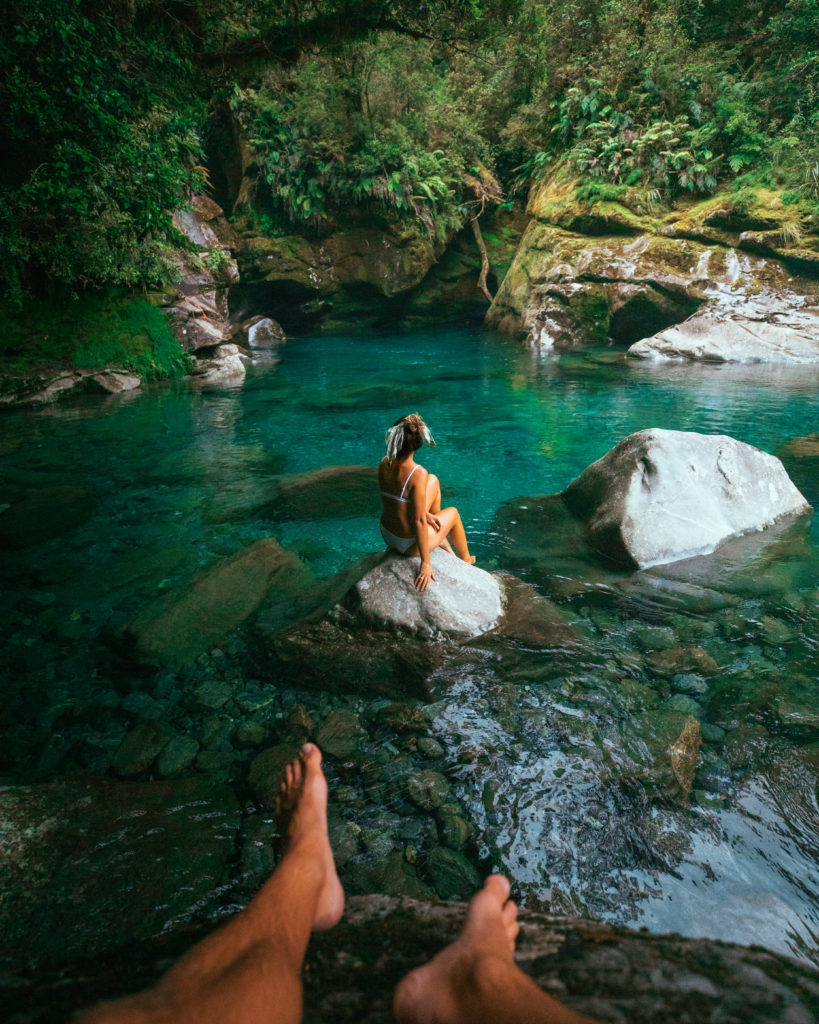 New Zealand Travel Guide | Point of view capture of woman sitting on a rock in a blue pool of water during travel adventure to New Zealand.