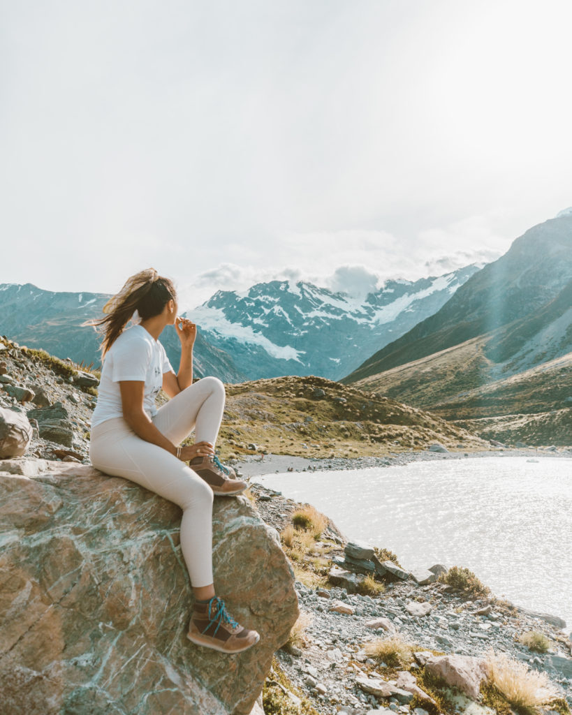 New Zealand Travel Guide | Woman on New Zealand travel adventure mindfully connects with nature as she sits on a rock, looking at the mountains of Mount Cook. Nearby lake glistens in New Zealand sunlight.
