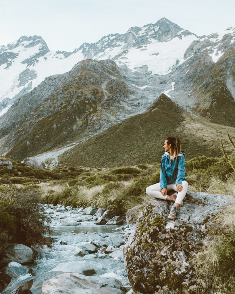 New Zealand Travel Guide | Woman wearing blue jacket sits in nature near mountains while hiking iconic Hooker Valley Track in Aoraki Mount Cook National Park in New Zealand. Small rock lined stream passes through green valley at the foot of the mountains.