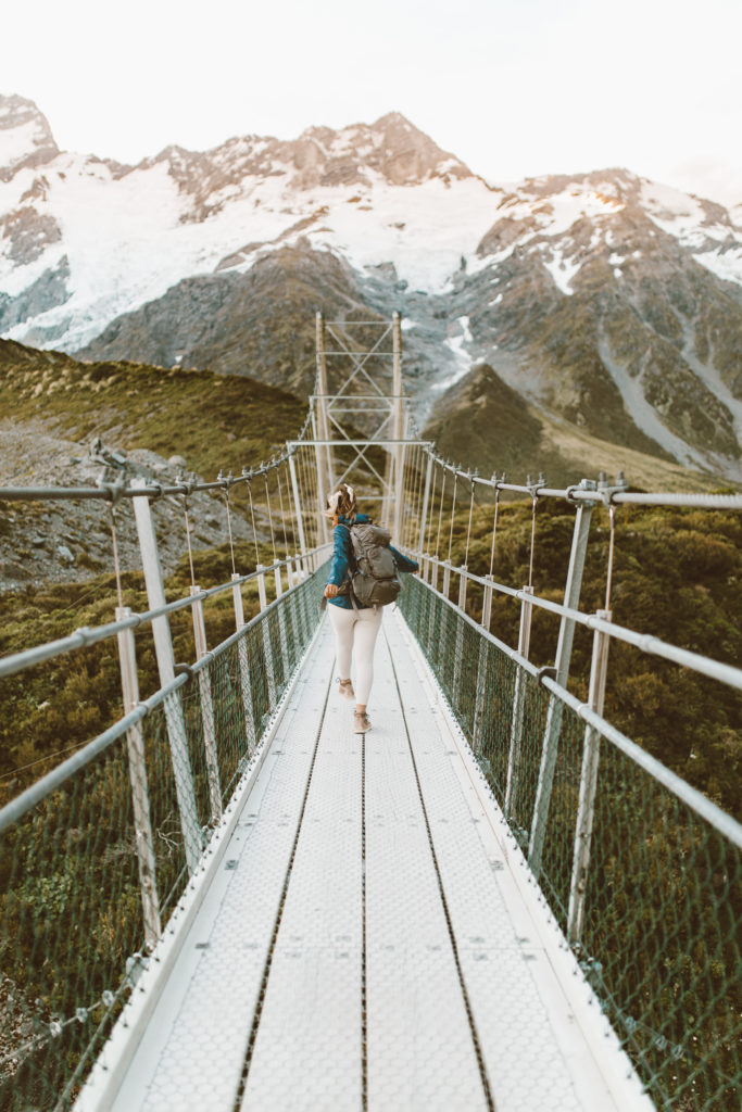 New Zealand Travel Guide | Hiker with backpack makes way across narrow bridge at iconic Hooker Valley Track in Aoraki Mount Cook National Park, in New Zealand. Tall snowy mountains await beyond the bridge.