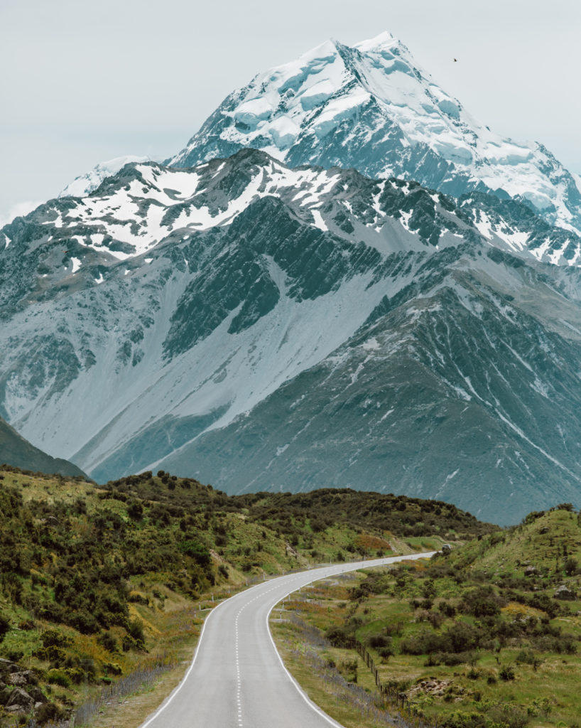 New Zealand Travel Guide | Open road takes a turn, leading to Mount Cook National Park. Large mountain spotted with snow stands tall against the green terrain, along the road, captured on trip to New Zealand.
