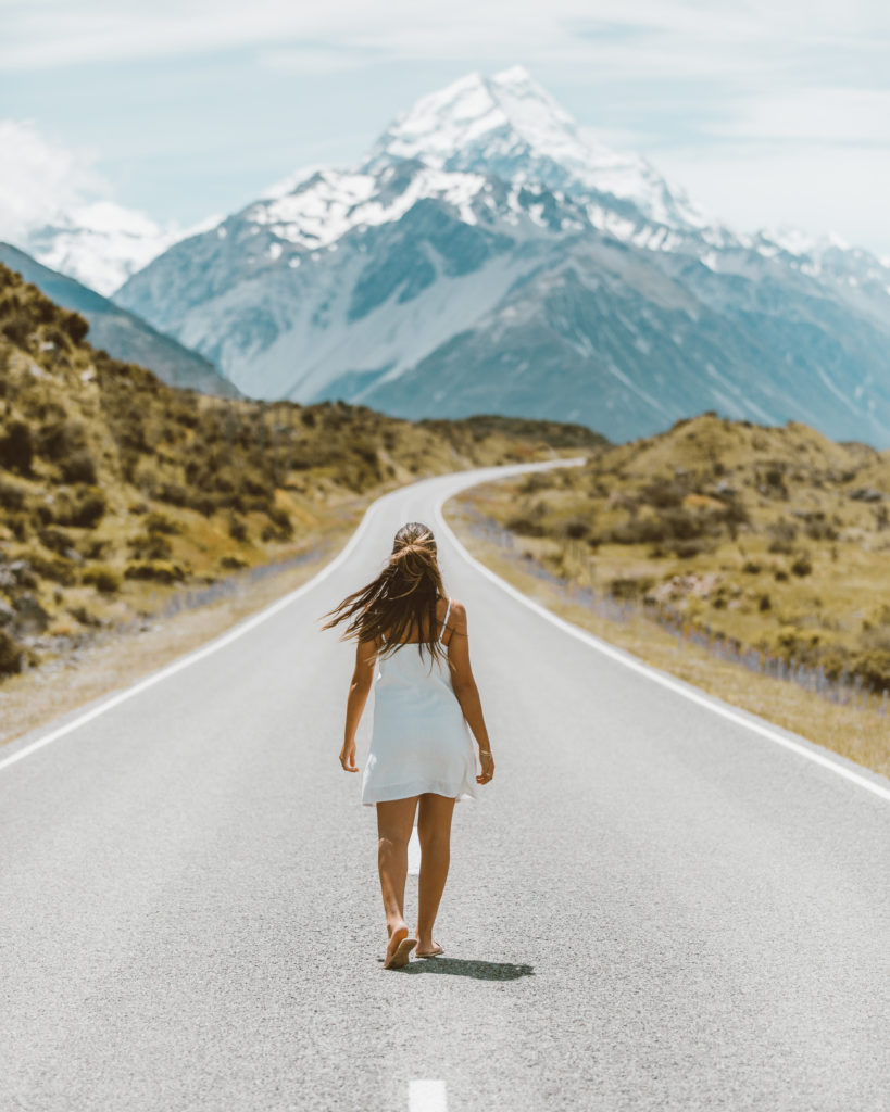New Zealand Travel Guide | Woman traveling on New Zealand adventure wearing a white dress and walking on road toward towering mountains located at Aoraki Mount Cook National Park. Mountain view shows mountains fading into soft haze in the distance.  