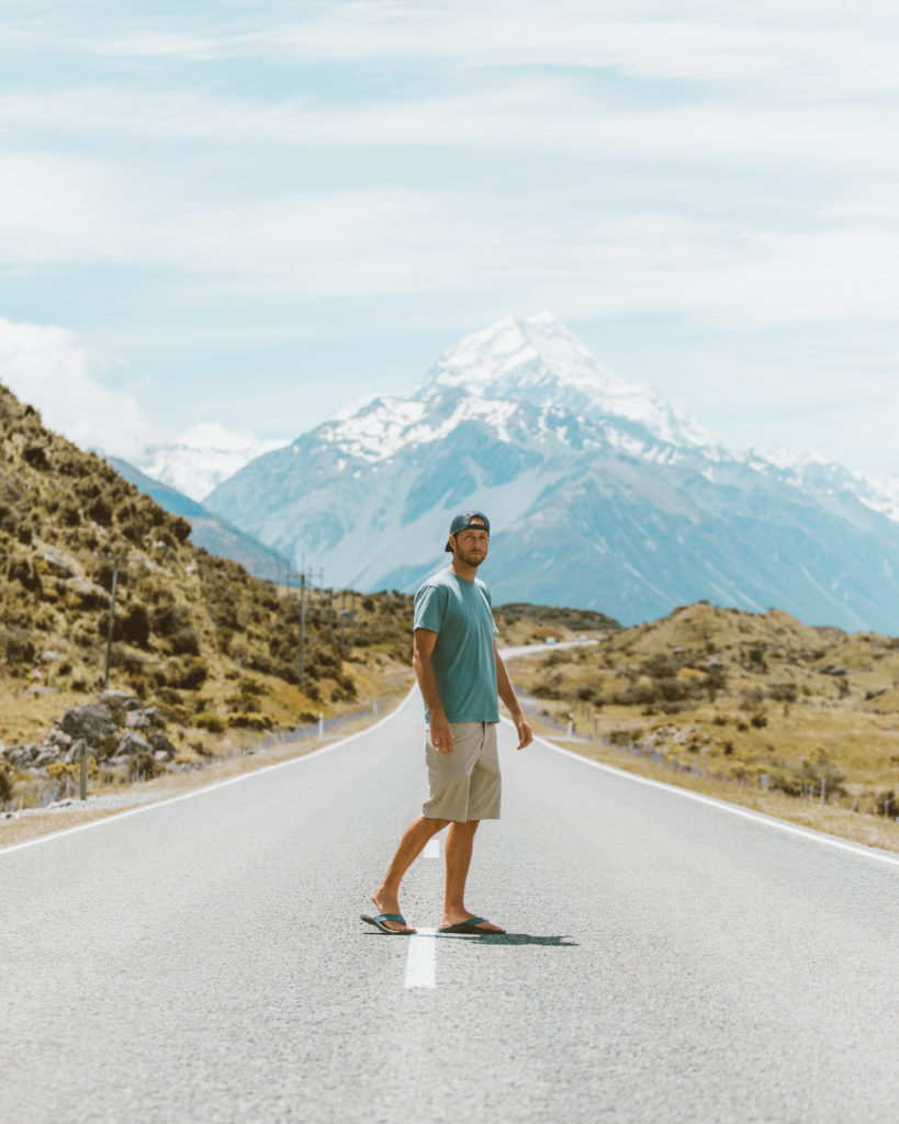 New Zealand Travel Guide | Man dressed in casual clothing, traveling to New Zealand, stands in a winding road between green grassy hills, with peaks of gorgeous Mount Cook mountain visible in the background