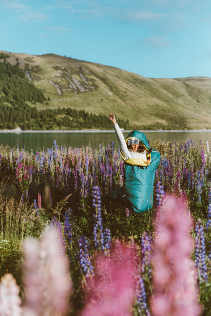 New Zealand Travel Guide | Woman in field of blooming lupins enjoys freedom camping near Lake Tekapo with L.L. Bean camping gear. Tall green hill and water in background, leading to a flower-filled field.