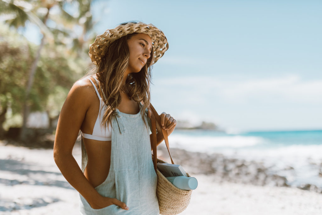 Woman on the beach holding with lauhala hat and overalls on. Ditch plastic water bottles when you travel with this portable disinfecting system. Sharing ways to reduce single-use plastic via @elanaloo + elanaloo.com